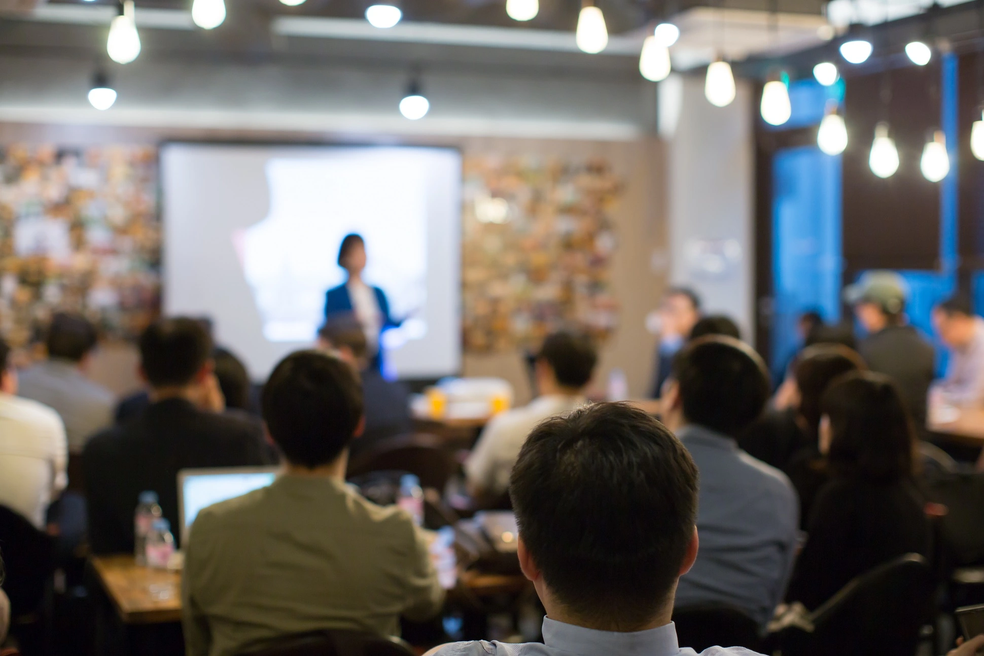 A woman leading a presentation with a projected image behind her. The camera is focused on the back of someone's head that is closer to the camera.